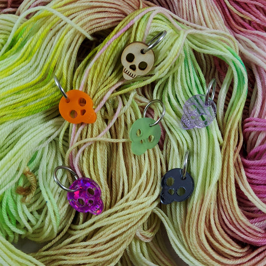 Six colorful skull shaped stitch markers on a background of colorful yarn