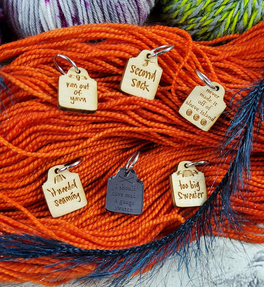 Six stitch markers shaped like tomb stones, engraved with the names of failed knitting projects.