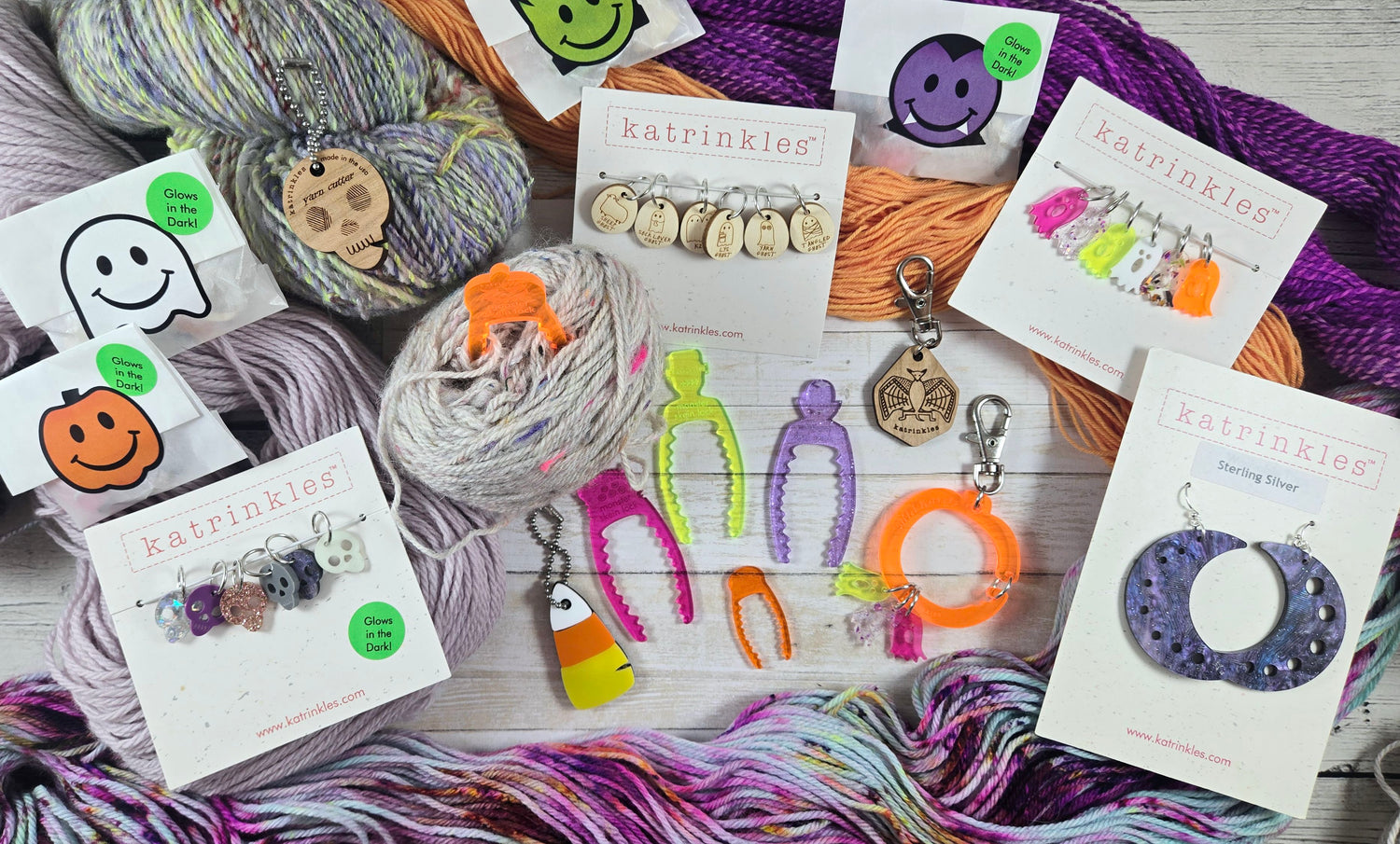 A flat-lay display of colorful yarn skeins and knitted accessory cards branded 'katrinkles' on a light wood background. The products include multicolor yarn, small knitted charms, metal clips, and circular knit pieces in purple, orange, pink, green, and gray tones.