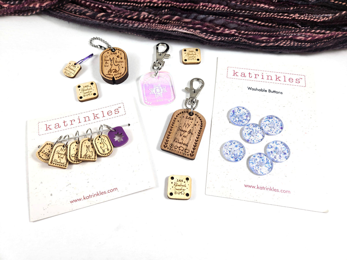 A flat-lay of KATRINKLES buttons and stitch markers, with a skein of blue yarn at the top. The set includes wooden and acrylic stitch markers, a cute yarn cutter, a Rhinebeck Bag Charm, an iridescent Woolen Affair bag charm, and blue-and-white glittery buttons on branded cards.
